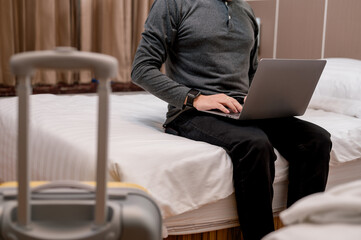 A man is sitting on a bed with a laptop in front of him. He is wearing a watch and has a suitcase...