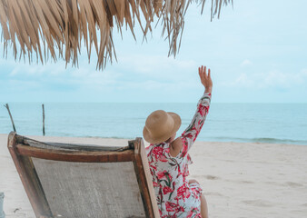 A woman is sitting on a beach chair with a hat on her head. She is waving at the camera
