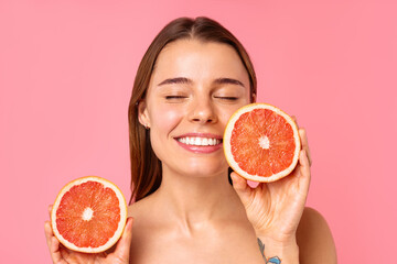 A woman smiles with grapefruit halves on pink background, symbolizing wellness and happiness