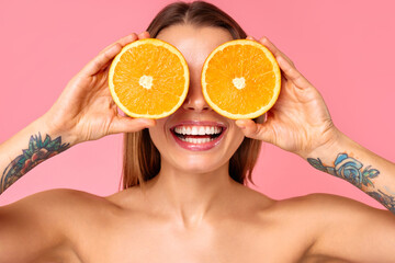 A joyful woman is smiling while wearing sliced oranges as glasses against a pink background