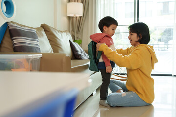 Asian Mother helps young son with green backpack stand in living room. Child in hoodie and leggings looks at mother while she adjusts backpack. Morning routine, preparation for school.