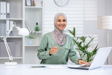 Confident businesswoman wearing hijab sitting at desk with laptop and glass of water, working in modern office. Professional career and productivity concept.