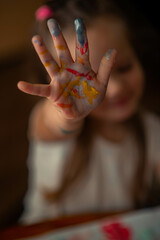 a 5-year-old girl with long blond hair, dressed in a light T-shirt, shows her hands stained with colorful paints, demonstrating her creativity