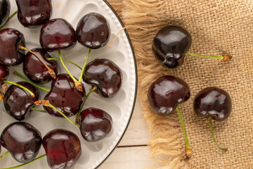 Several ripe sweet cherries with ceramic saucer and jute napkin on wooden table, macro, top view.