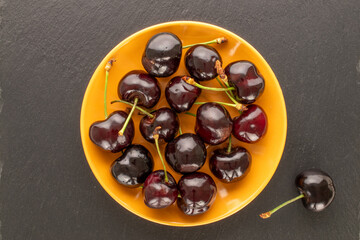 Several ripe sweet cherries with ceramic saucer on slate stone, macro, top view.
