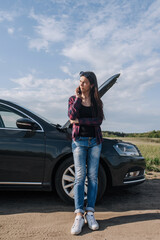 A young beautiful lonely woman driver stands near a broken down black car with an open hood on the...