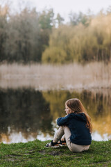 Little beautiful red-haired girl, lonely dreaming child sits on green grass against the backdrop of...