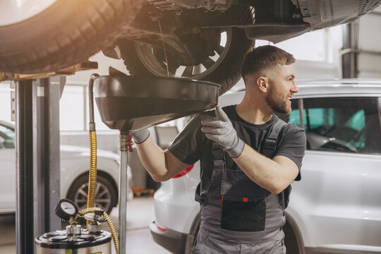 A car workshop worker is draining oil from engine while standing under the car
