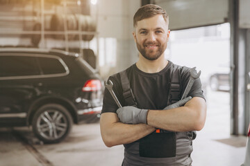 Smiling mechanic looking at camera while standing with crossed arms and holding wrenches in a car service