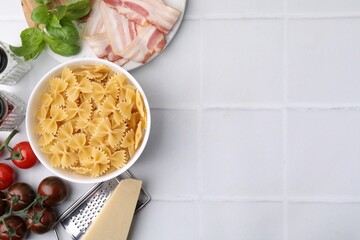 Raw pasta, bacon and fresh ingredients on white tiled table, flat lay. Space for text