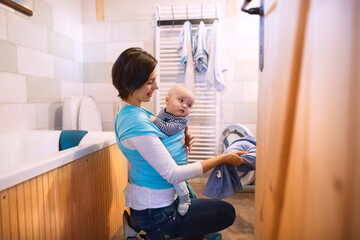 Mother holding small baby while loading the washing machine with dirty laundry, carrying him around...