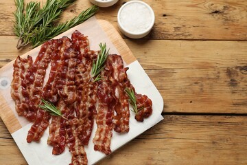 Slices of tasty fried bacon, rosemary and salt on wooden table, top view, space for text