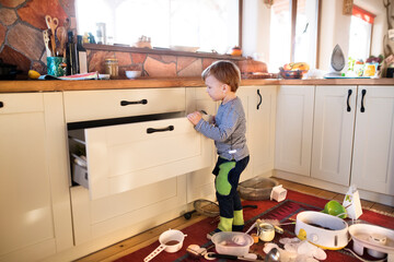 Little boy is taking kitchen utensils out of the drawers, making a mess in the kitchen.