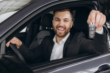 Cheerful bearded man holding key from auto, buying brand new car in showroom. Happy smiling guy...