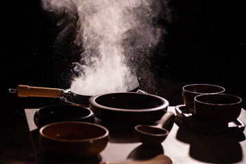 The cook was using a spatula to fry food in a pan of hot oil. There was a lot of smoke rising from the pan. There are wooden cups and plates placed on the table in a retro style.