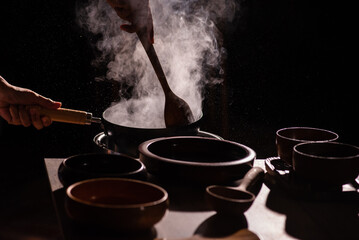 The cook was using a spatula to fry food in a pan of hot oil. There was a lot of smoke rising from the pan. There are wooden cups and plates placed on the table in a retro style.