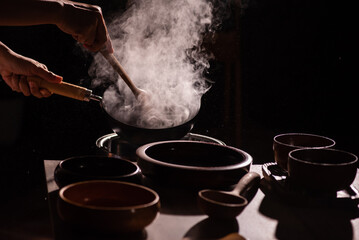 The cook was using a spatula to fry food in a pan of hot oil. There was a lot of smoke rising from the pan. There are wooden cups and plates placed on the table in a retro style.