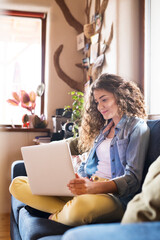 A beautiful university student having online class, e-learning sitting at home on the couch, laptop...