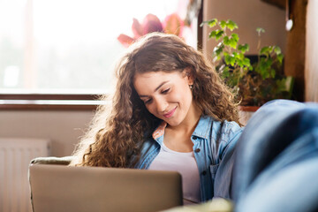 A beautiful university student having online class, e-learning sitting at home on the couch, laptop...