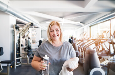 Overweight woman resting after workout in gym. Drinking water from bottle.