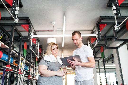 Overweight woman consulting with trainer, discussing daily workout plan. Personal trainer talking with client, discussing exercise program in gym.