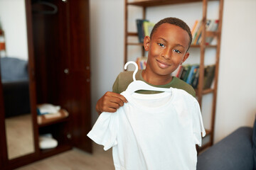 Happy boy kid with dark skin holding clothes-hanger with white t-shirt standing in his room, satisfied with his choice and purchase of new outfit for training workouts and playing outdoors