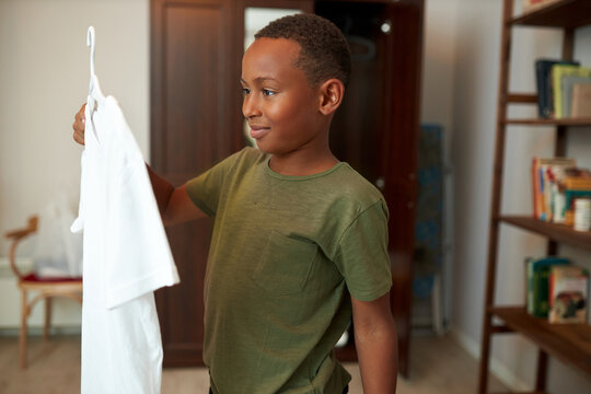 Side view of happy little african american boy preparing for party or event at school standing in his room holding clothes-hanger with white t-shirt, looking with anticipation to wear it asap