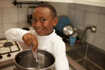 Excited happy black boy kid in white clothes holding cooking pot and whisk pretending to whip cream standing in stylish kitchen with big round eyes, making cake for first time to surprise his mom