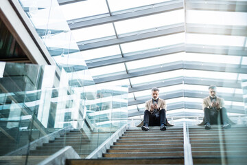 Handsome businessman sitting on stairs in modern office hallway, scrolling on smartphone.