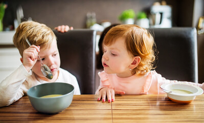 Siblings sitting at table, eating lunch, green soup. Girl looking in brother's plate.