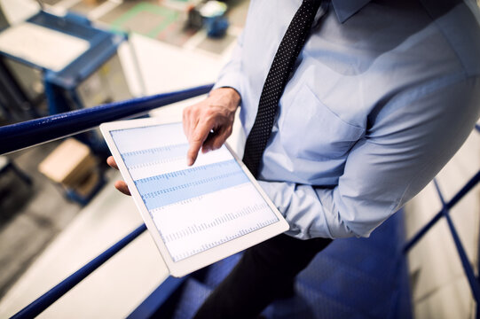 Manager standing in modern industrial factory, scrolling on tablet. Manufacturing facility with robotics, robotic arms and automation. Storing products and materials in warehouse.