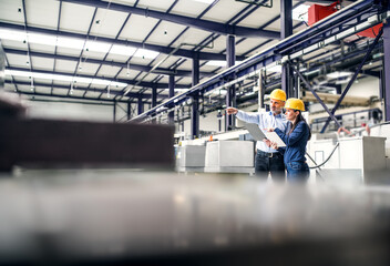 Two project managers standing in modern industrial factory. Manufacturing facility with robotics,...