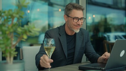 Mature businessman in modern cafe, smiling and holding champagne glass, working on his laptop...