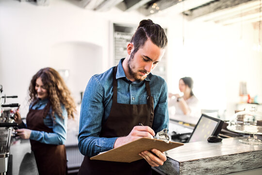 Two young baristas working in coffee shop, standing by counter. University students working part-time in cafe.