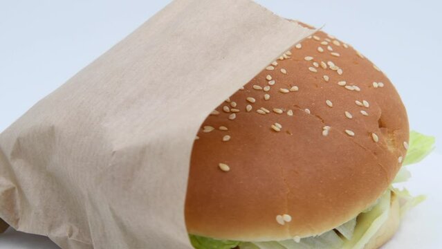 Close-up view of chickenburger with letuce salad in paper bag spinning against gray background. Bun covered with sesame seeds. Soft focus. Real time video. Fast food restaurant. Unhealthy eating theme