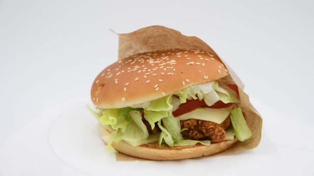 Close-up view of chickenburger with letuce salad in paper bag spinning against gray background. Bun covered with sesame seeds. Soft focus. Real time video. Fast food restaurant. Unhealthy eating theme