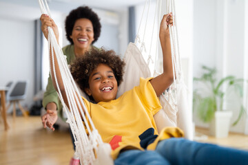 African American young girl playing on a rope swing in the living room at home. Mother swinging a...