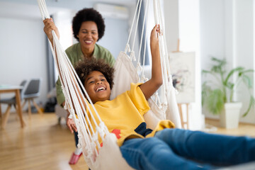 Cheerful African American young girl playing on a rope swing in the living room at home....