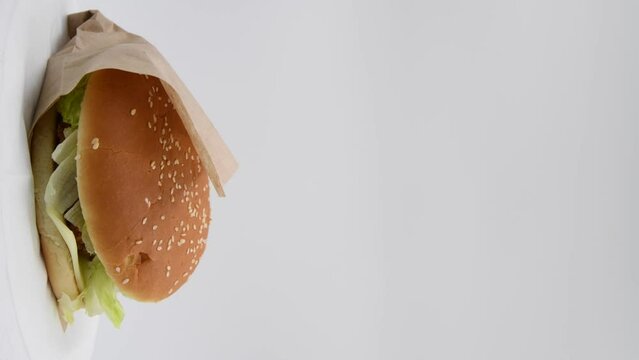 Close-up view of chickenburger with letuce salad in paper bag spinning against gray background. Bun covered with sesame seeds. Vertical video. Copy space. Fast food restaurant. Unhealthy eating theme.