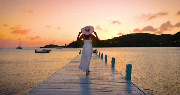A beautiful woman in a white dress and with hat enjoys the tropical sunset in the Caribbean Sea, Antigua and Barbuda island