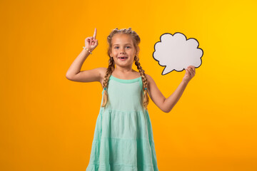 Thoughtful kid girl holding cloud bubble card and pointing finger up over yellow background. Idea concept