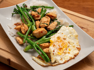 Asian dish with fried chicken, broccolini, fried egg and wide noodles served on a white plate on a wooden board.