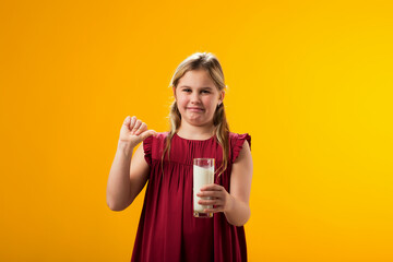 Obraz premium Kid girl with dairy allergy holding glass of milk and showing thumb down gesture on yellow background. Lactose intolerance concept