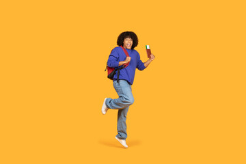 A cheerful guy wearing a blue sweater and carrying a red backpack jumps in excitement while holding a passport and ticket. The vibrant orange background adds energy and optimism to the scene.