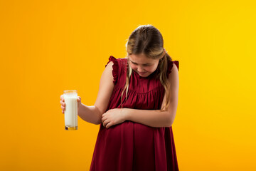 Kid girl holding glass of milk and feeling abdominal pain on yellow background. Lactose intolerance...