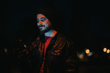 Man wearing a leather jacket using his smartphone at night, illuminated by the screen with city...