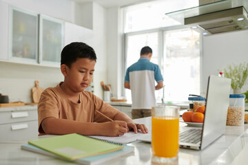 Vietnamese boy doing homework at kitchen counter when father cooking lunch