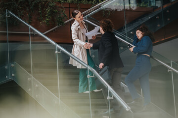 Three businesspeople greeting and exchanging documents on a staircase in a modern office setting....