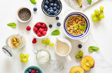 Set of various ingredients for cooking a healthy breakfast setup with granola, assorted fresh fruits, chia seeds and yogurt on a white marble background top view