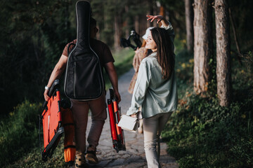 Carefree friends walking on a forest path carrying musical instruments and camping gear,...